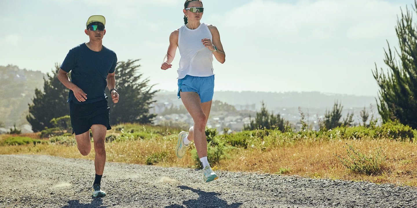 Man in navy tee and woman in white tank and blue shorts running on a gravel path with coastal hills behind