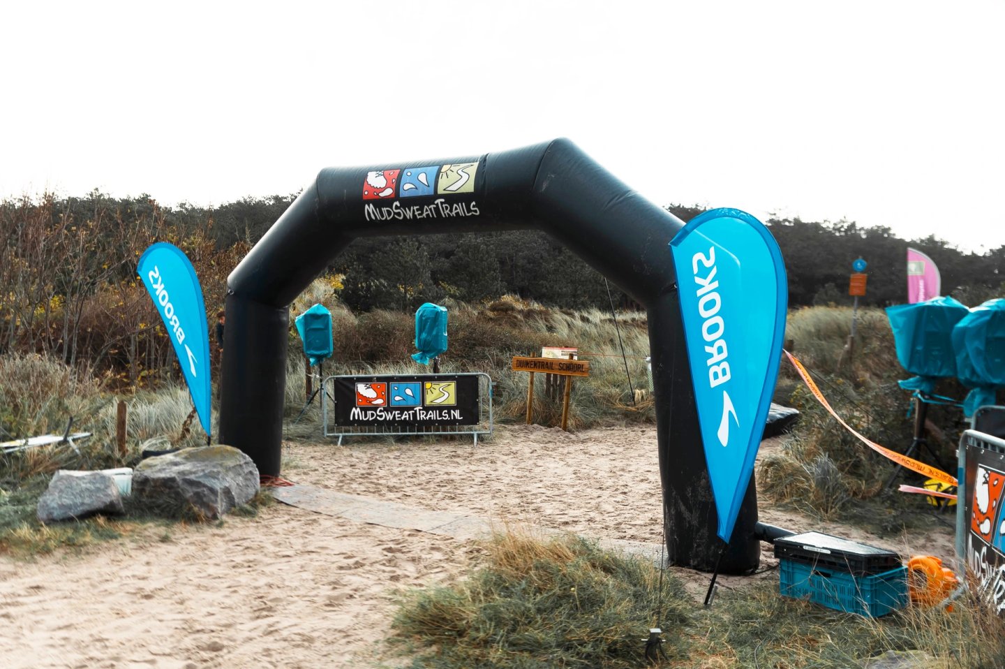 Black inflatable MudSweatTrails race arch with Brooks teardrop flags on sandy dune terrain on an overcast day