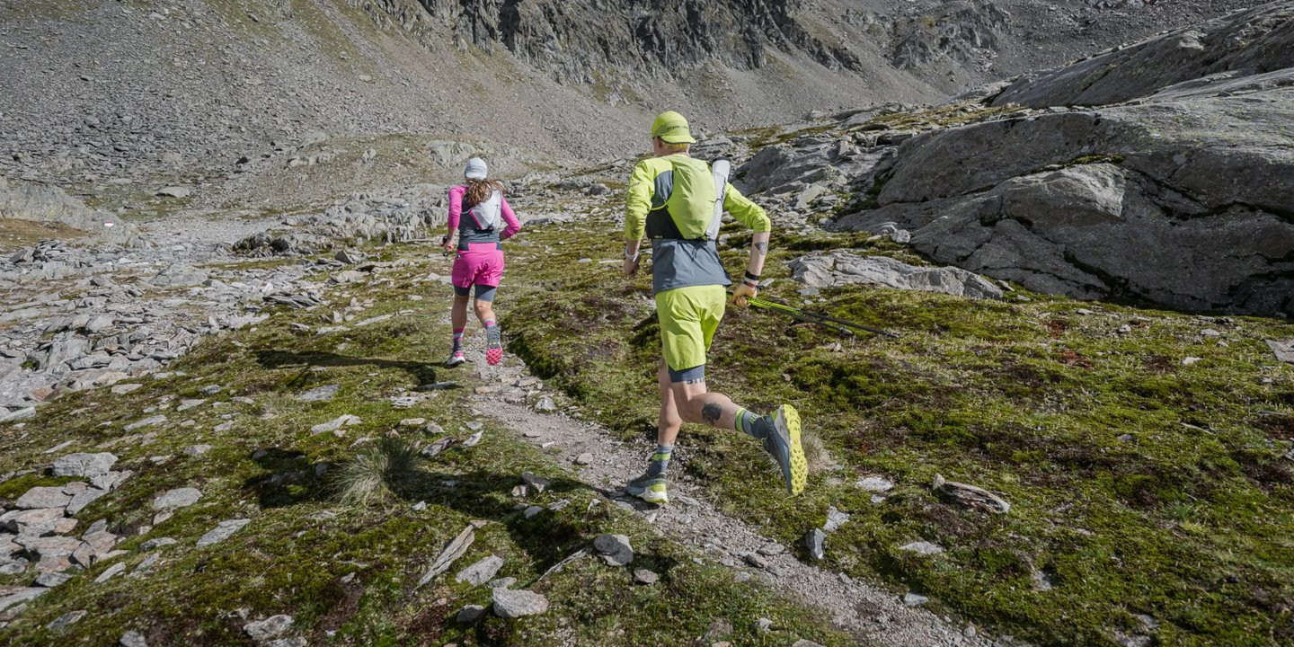Two trail runners in bright sports kit ascending a rocky alpine mountain path with trekking poles and hydration vests.