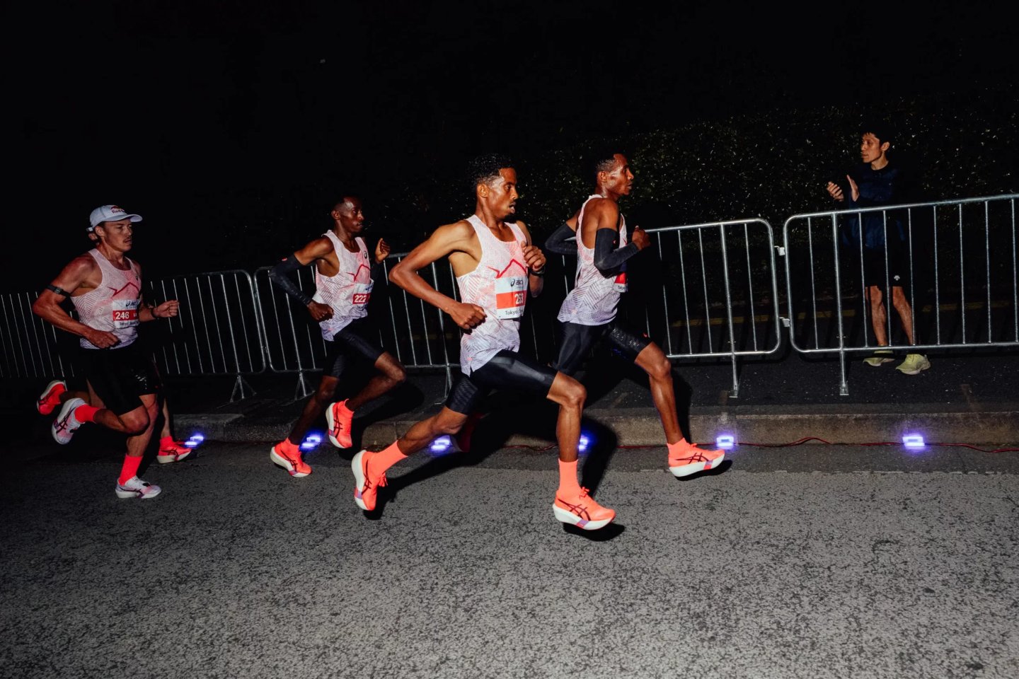 Four elite marathon runners in ASICS Metaspeed coral racing shoes competing in a night road race.