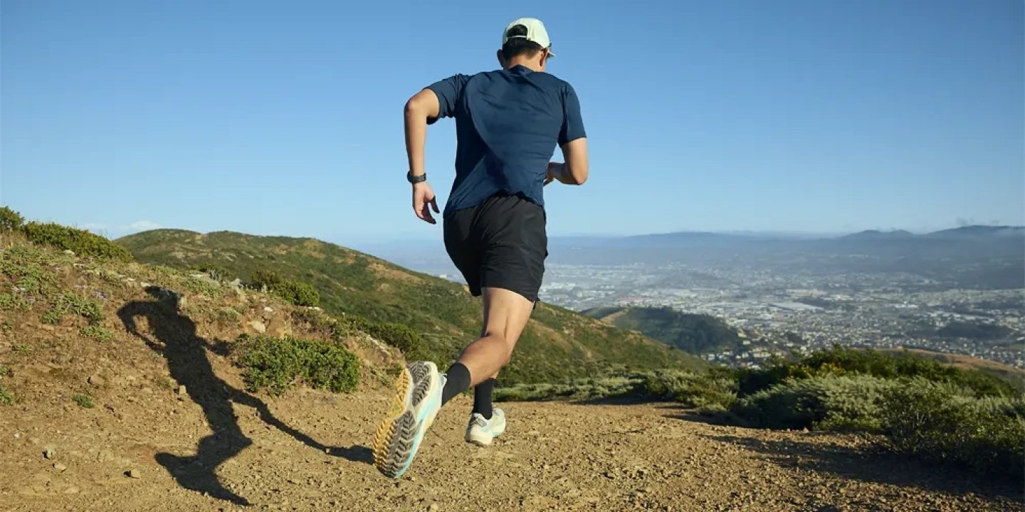 Male runner ascending a rocky dirt trail on a mountain ridge wearing blue shirt, black shorts and trail running shoes