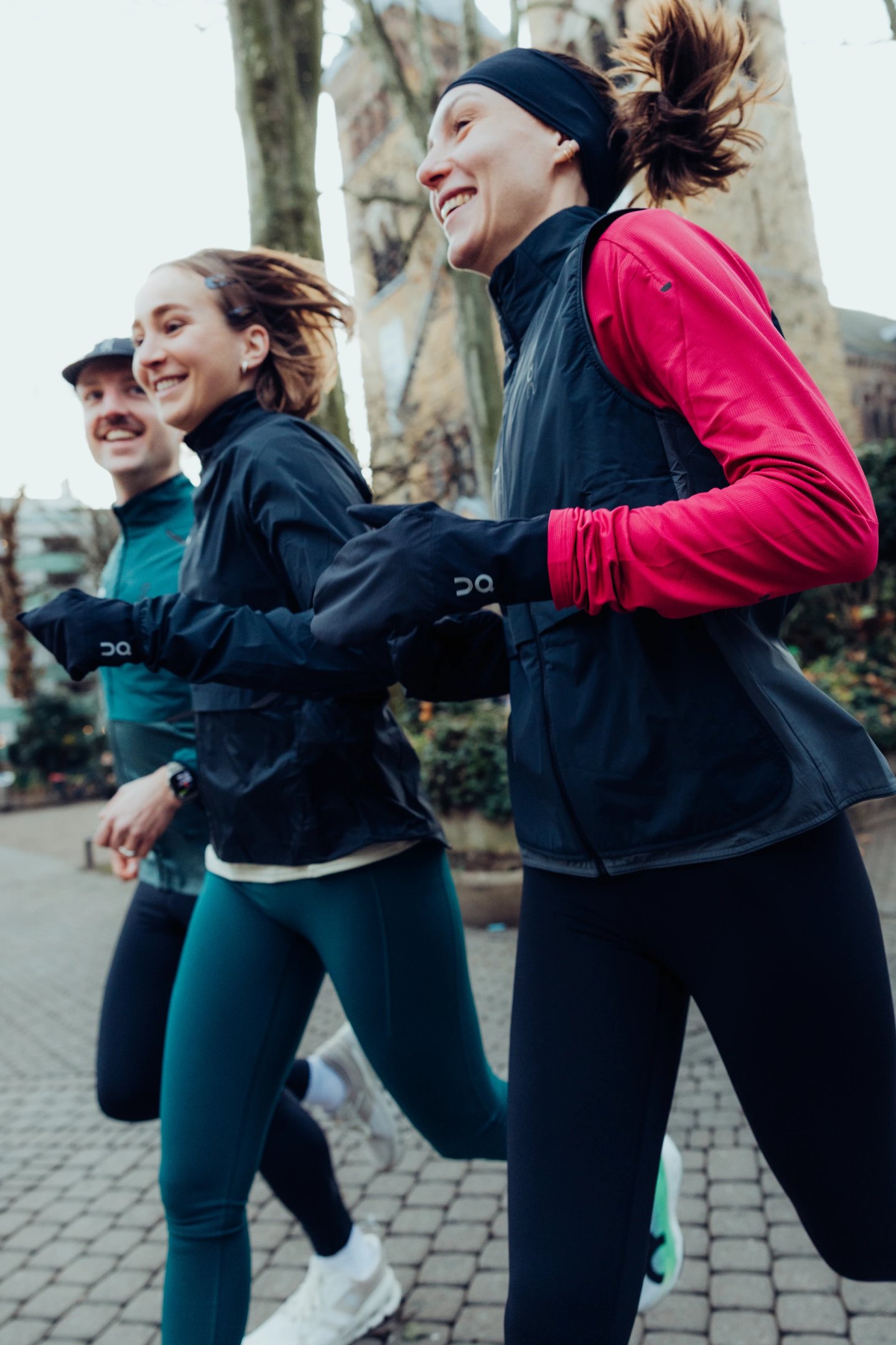 Three runners smiling while running on a cobblestone path in winter, wearing black running jackets, tights, and a pink long-sleeve top.