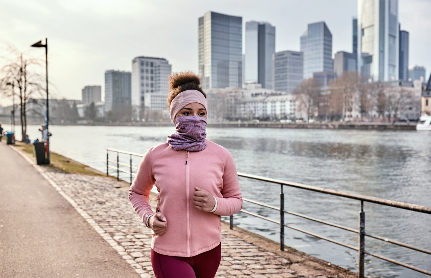 Woman running along a riverside city path in a pink fleece zip jacket, pink headband, and purple neck gaiter on a cold day.