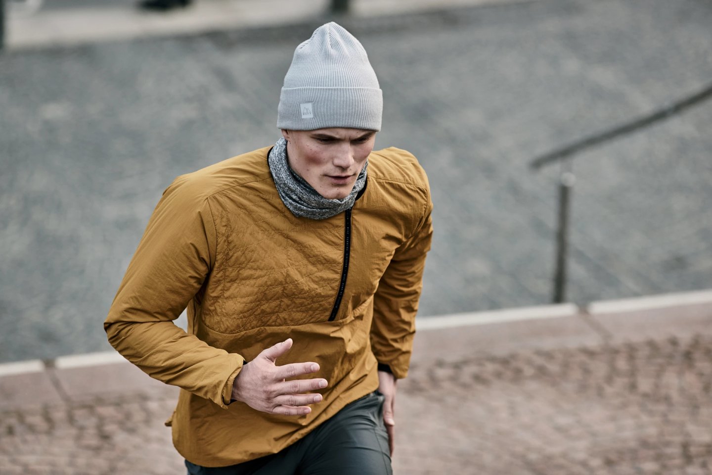 Male runner wearing a light grey City Running Crossknit beanie, mustard quilted jacket and neck gaiter, running outdoors in cold weather.