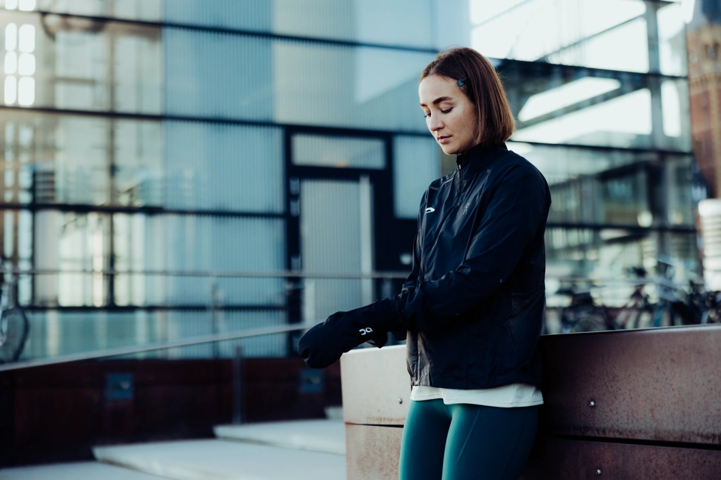 Woman in black running jacket, teal tights and black running gloves checking her gear on a city bench