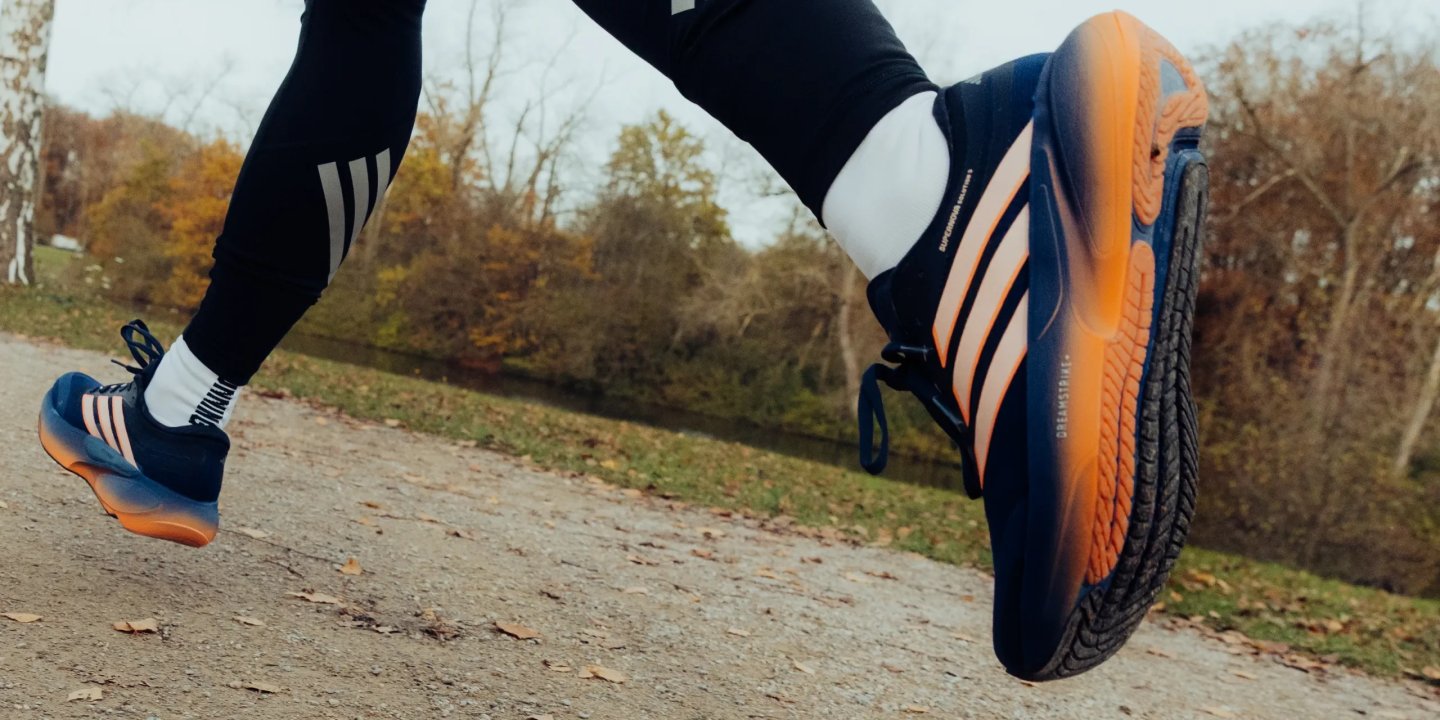 Runner mid-stride in Adidas Supernova Solution navy and orange running shoes on an autumn gravel path.