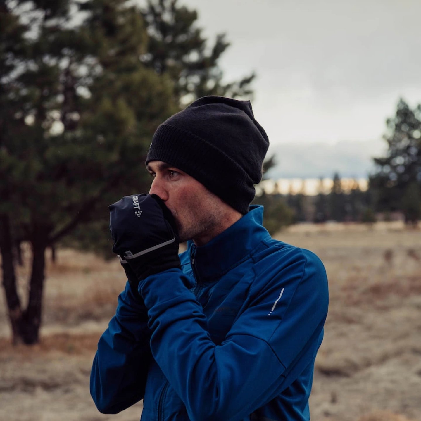 Male runner wearing Craft SubZ jacket in blue with black beanie and Craft running gloves in cold outdoor winter conditions.