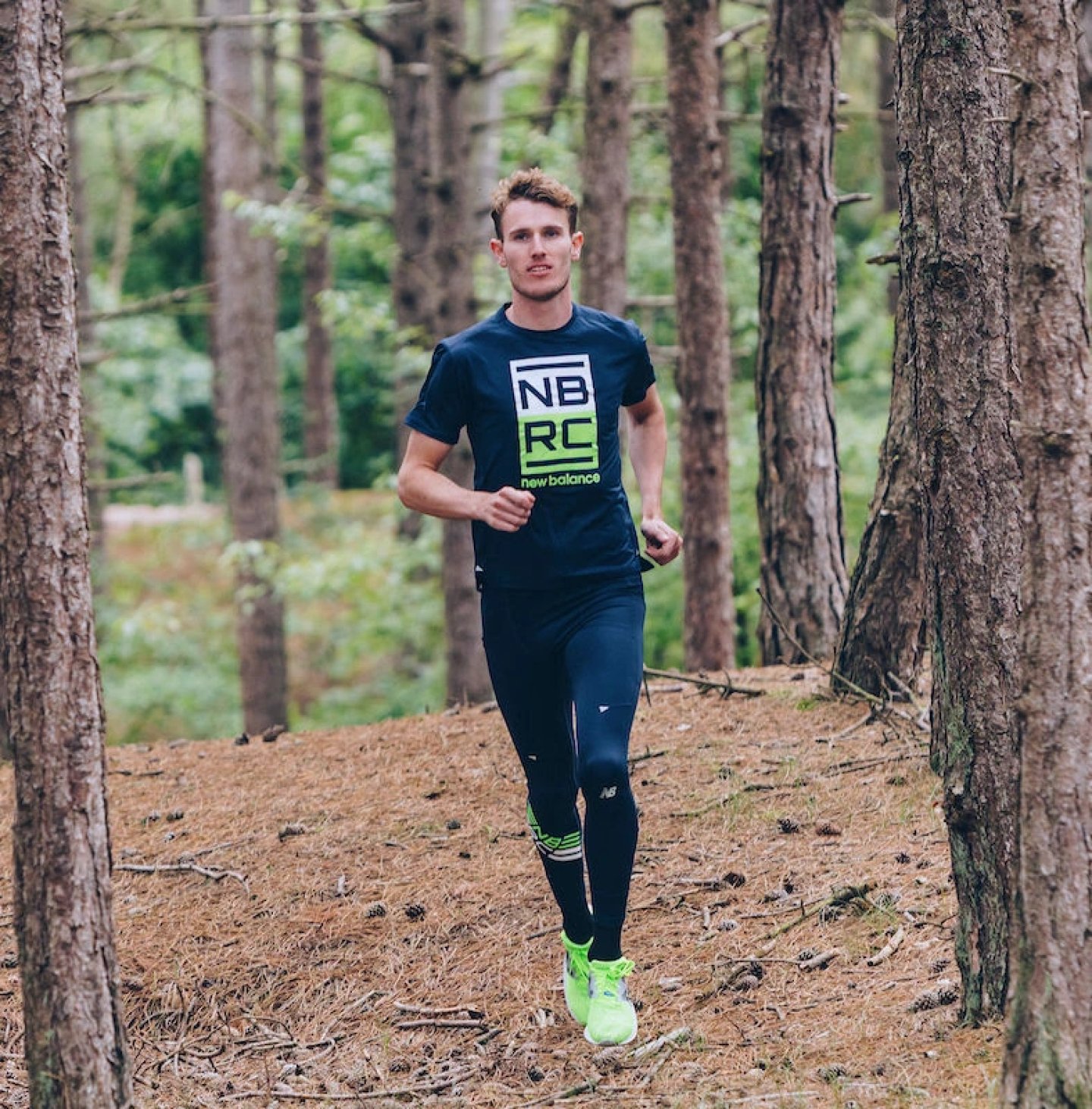 Male runner wearing New Balance NBRC navy graphic t-shirt, dark tights and neon yellow trainers running through a pine forest.